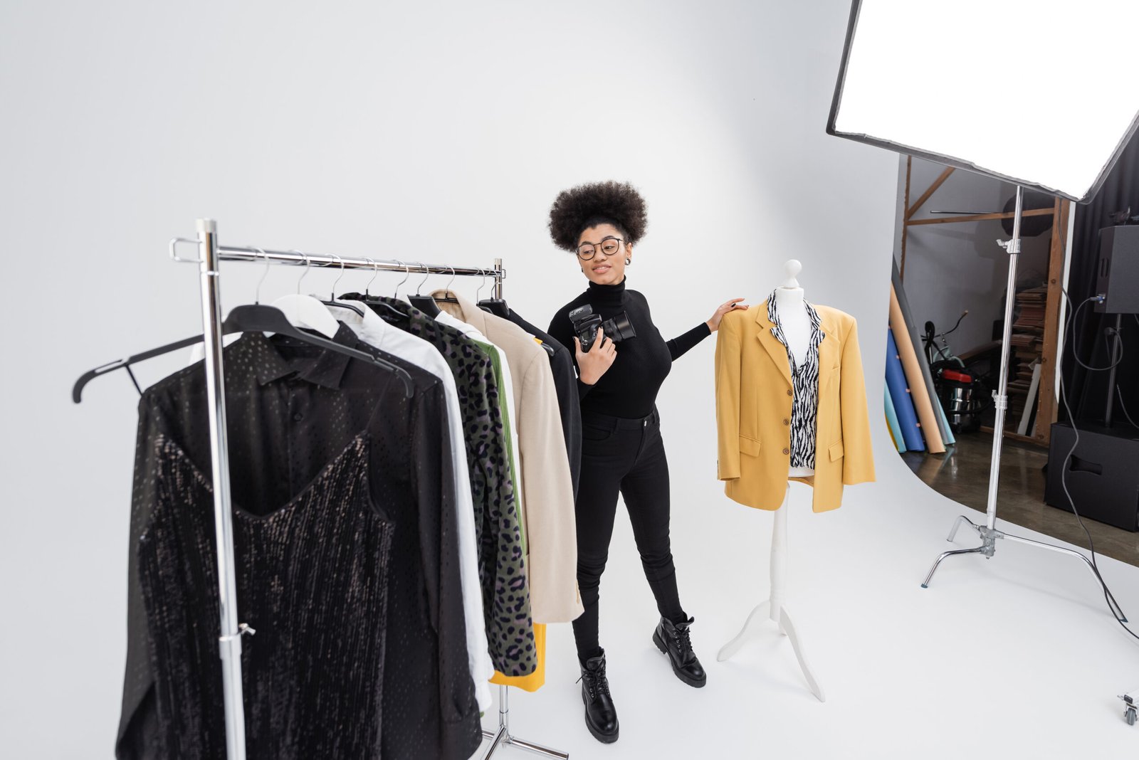full length of african american content maker with digital camera smiling near rail rack and mannequin with trendy clothes in photo studio